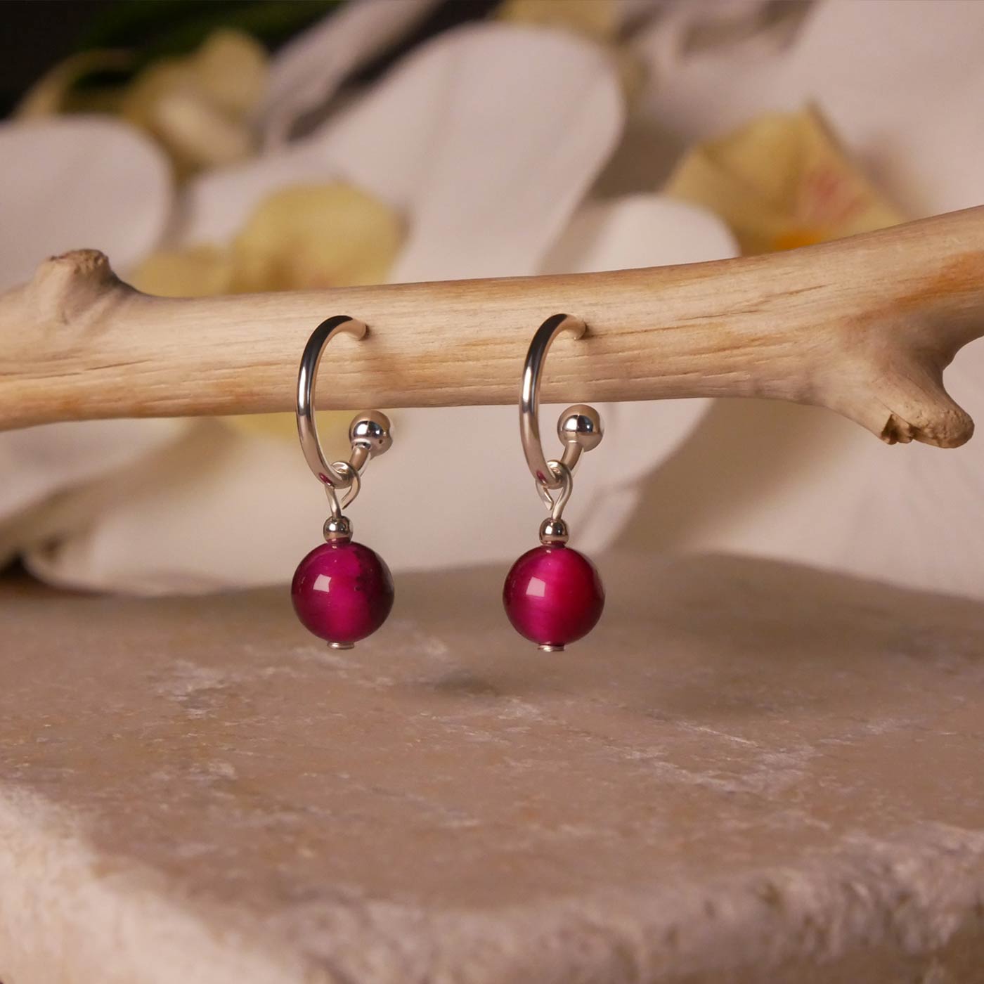 Sterling silver hoop earrings with dark pink tiger's eye round sphere charms hanging on a branch. It is stylized hanging over natural stone and with a blurred background of white orchids flowers.