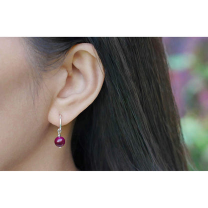 Close-up of a person's ear wearing a sterling silver hoop earring, with a dark pink tiger's eye round sphere charm. Person is outside, has long black hair, with the floral and plant background blurred.