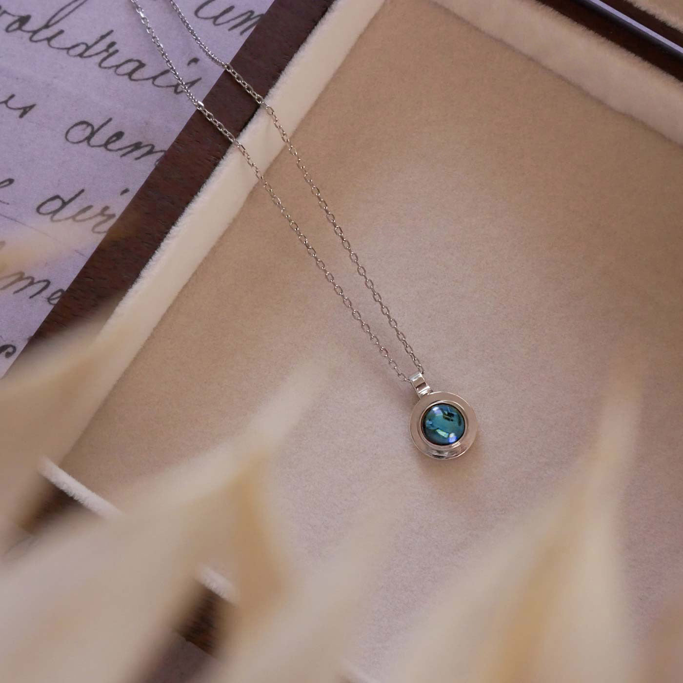 Top view of a dainty, round Azotic blue topaz cabochon pendant sterling silver necklace. It is stylized in a beige cloth lined wooden jewelry box with beige leaves in the foreground. The jewelry box sits on vintage paper underneath with cursive writing on it.