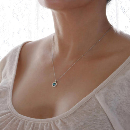3/4 view of a dainty, round Azotic blue topaz cabochon pendant sterling silver necklace, worn by a person wearing a light beige shirt.