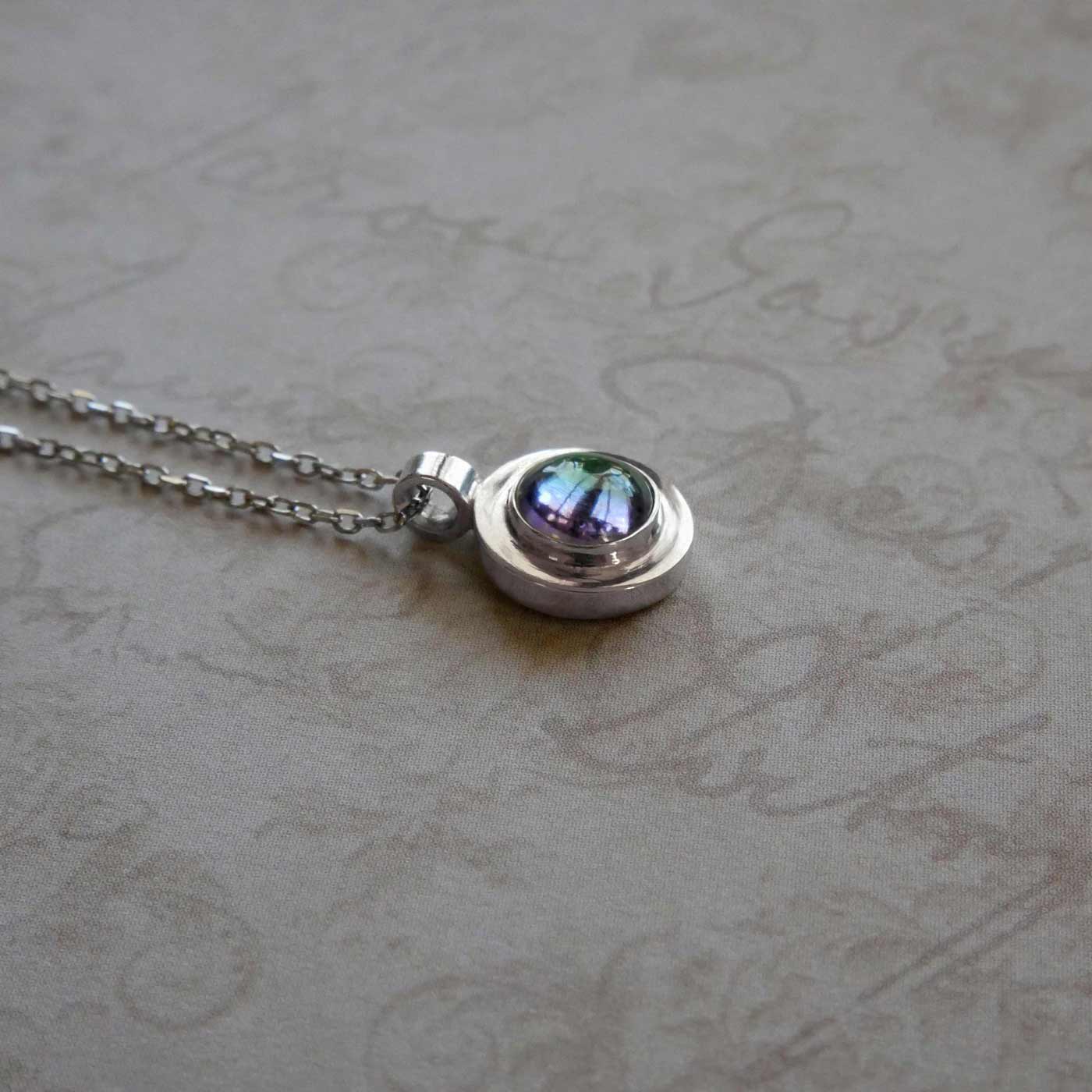 Close up 3/4 view of a dainty, round Azotic blue topaz cabochon pendant sterling silver necklace. It lays flat on a piece of vintage textured paper with cursive writing on it.