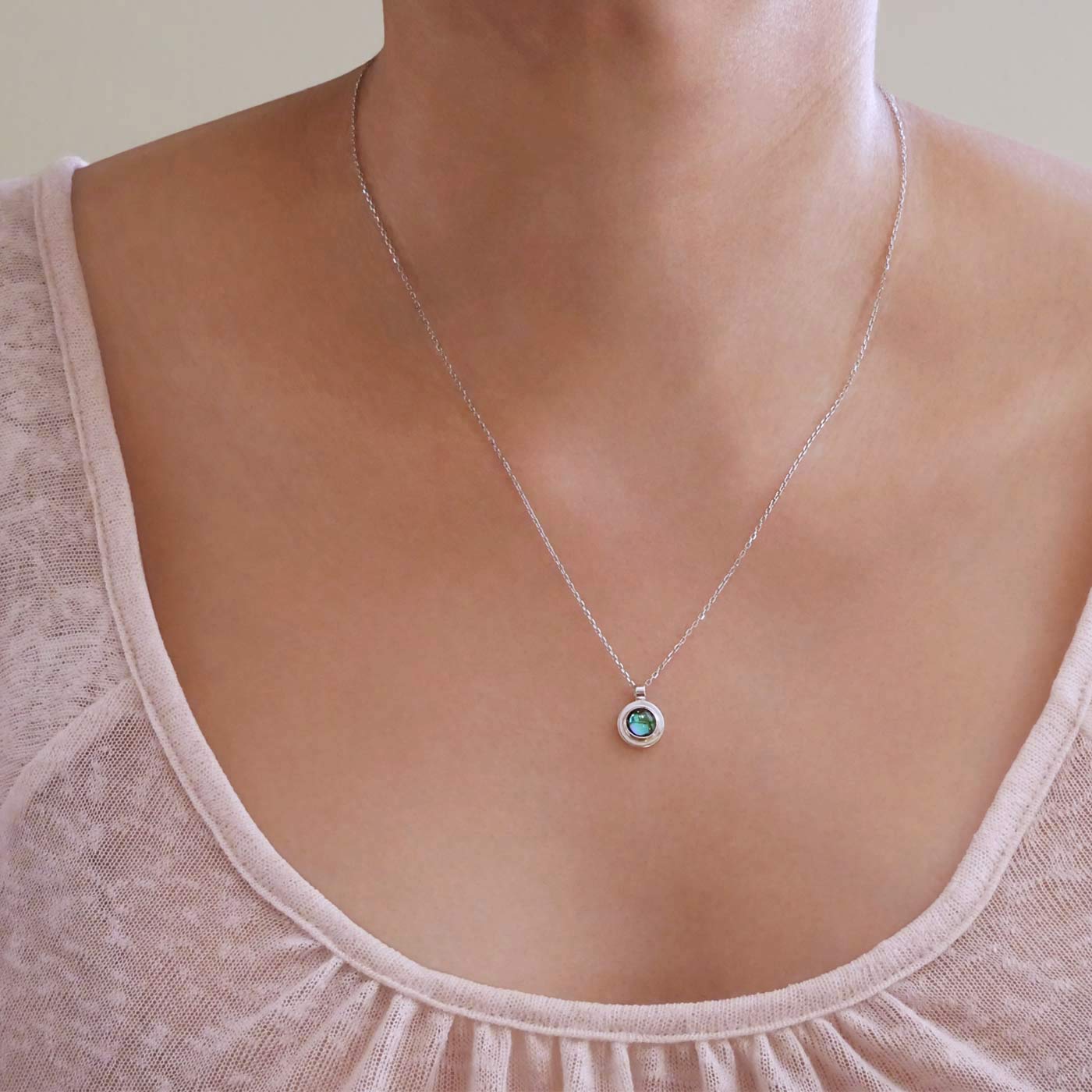 Front view of a dainty, round Azotic blue topaz cabochon pendant sterling silver necklace, worn by a person wearing a light beige shirt.
