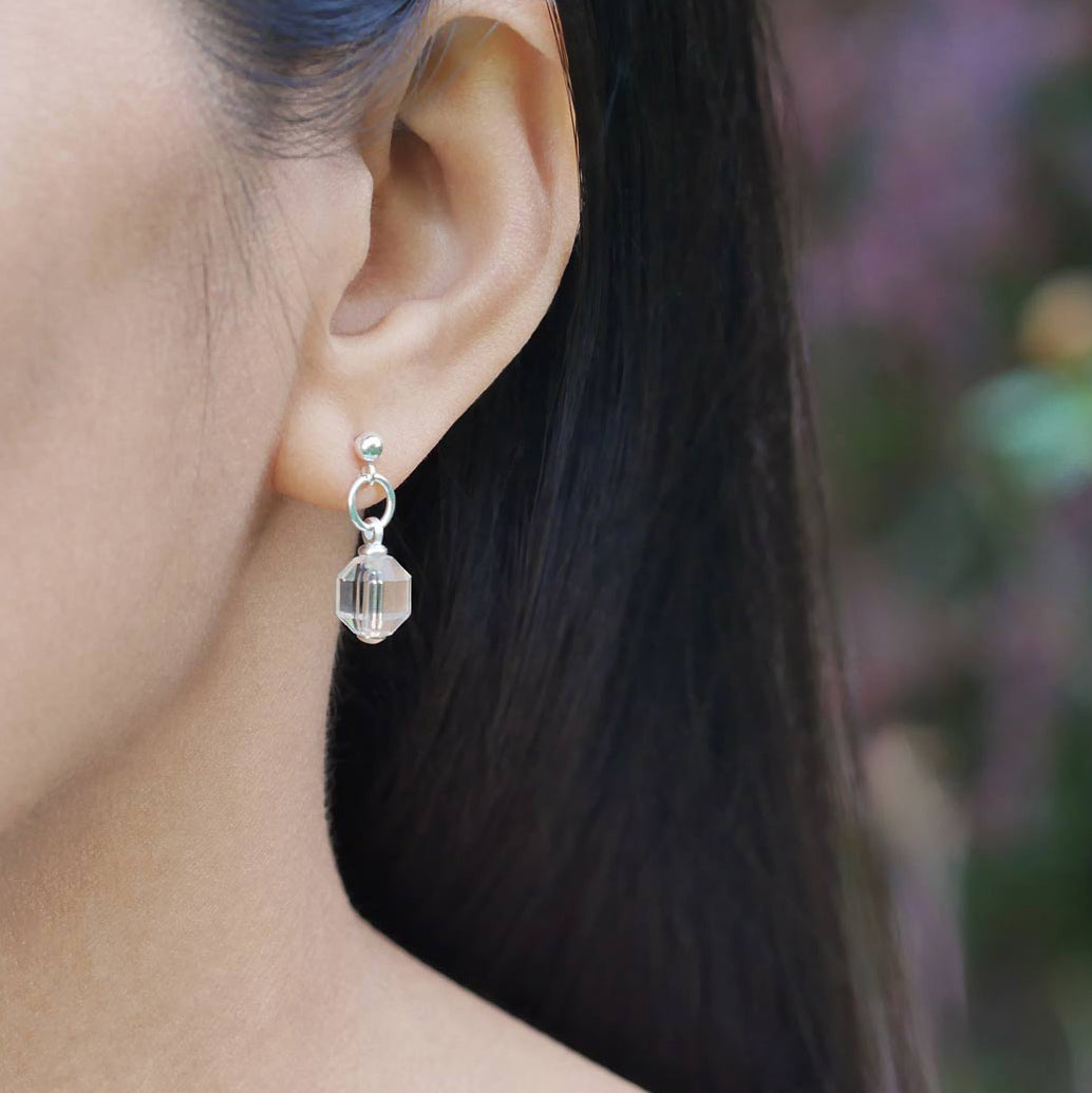 A close up of a sterling silver clear quartz crystal prism earring worn outside by a person with dark hair, against a blurred natural background with plants and flowers.