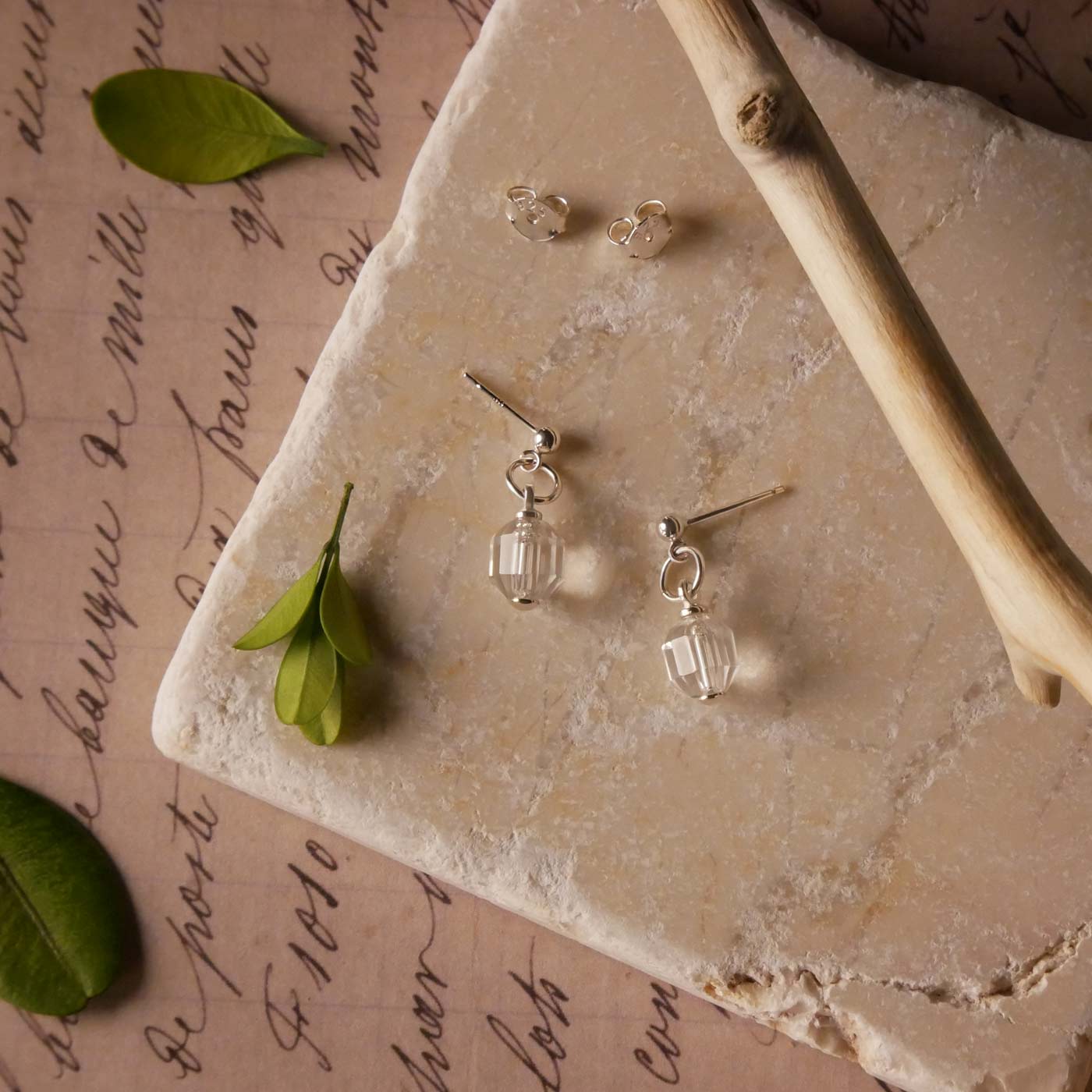 A pair of sterling silver earrings with clear quartz crystal prisms, laying flat on a textured stone surface. It is stylized with accent green leaves, a tree branch, and vintage paper with cursive writing on it.