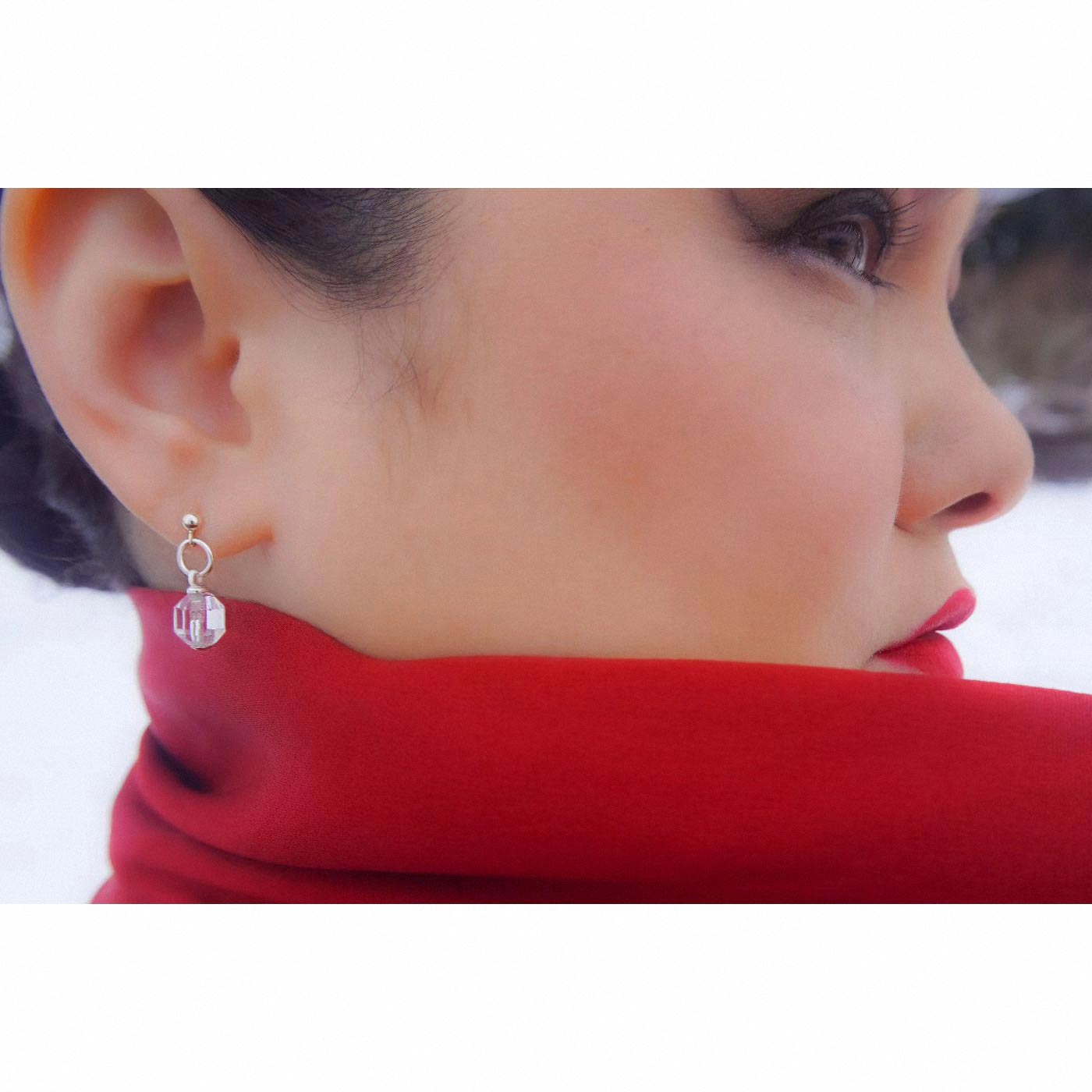 A close up of a sterling silver clear quartz crystal prism earring worn outside by a person with dark hair, wearing a red garment outside with white snow blurred in the background.