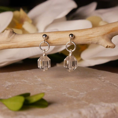 A pair of sterling silver earrings with clear quartz crystal prisms, hanging on tree branch over a textured stone surface. It is stylized with accent green leaves and white orchid flowers blurred  the background.