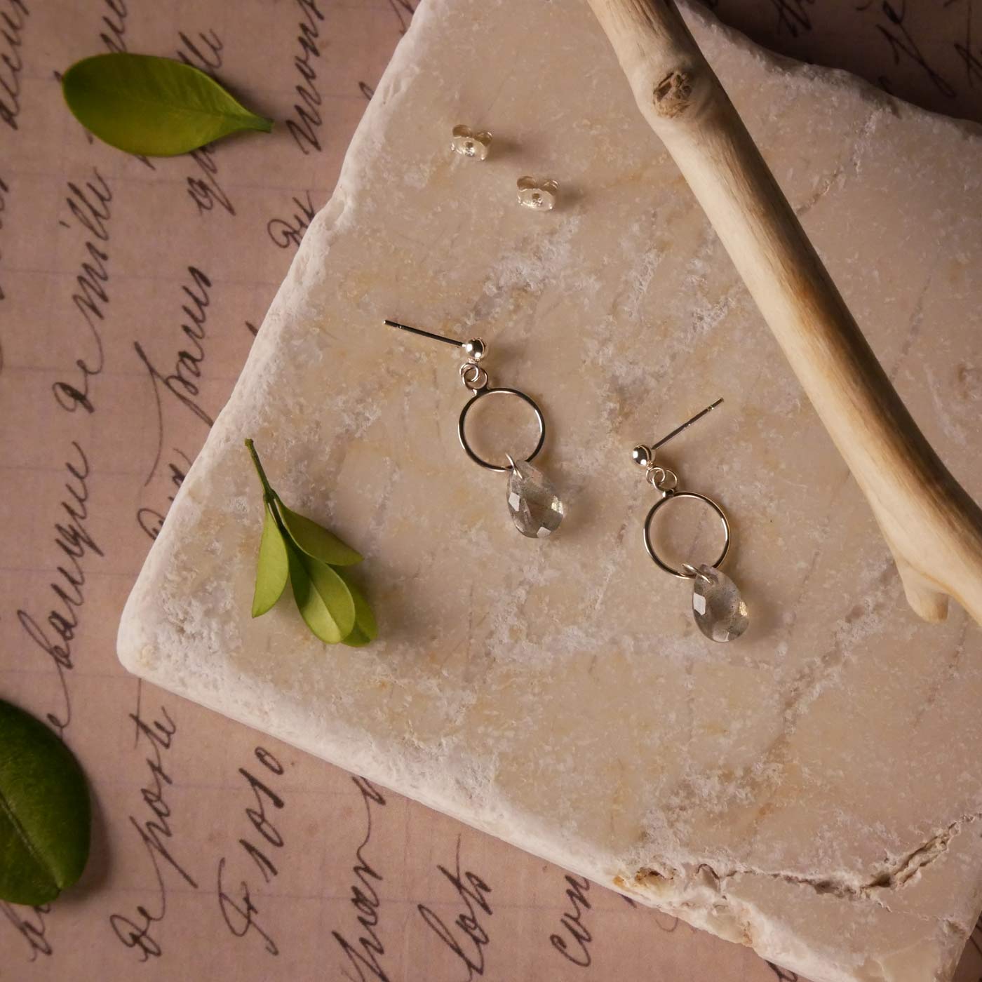 Top view of a pair of sterling silver, open circle labradorite drop earrings. It is stylized by laying flat on a natural stone surface, over vintage paper with cursive writing, and surrounded by green leaves with a branch in the foreground.