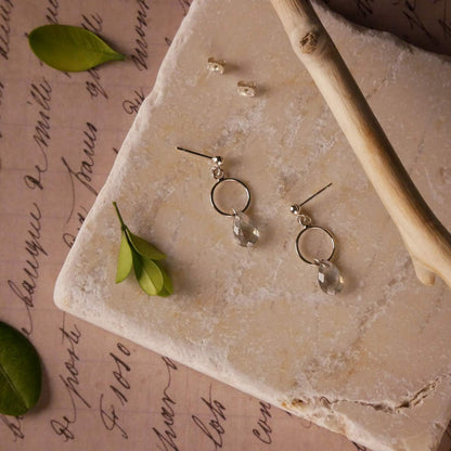 Top view of a pair of sterling silver, open circle labradorite drop earrings. It is stylized by laying flat on a natural stone surface, over vintage paper with cursive writing, and surrounded by green leaves with a branch in the foreground.