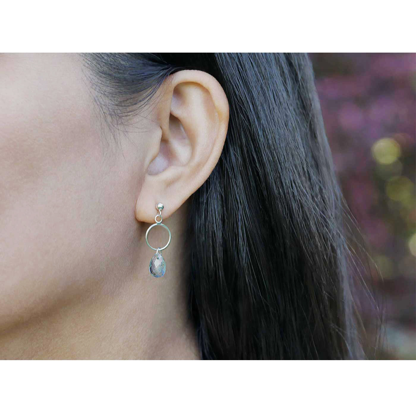 A close up of a sterling silver open circle labradorite earring worn outside by a person with dark hair, against a blurred natural background with plants and flowers.