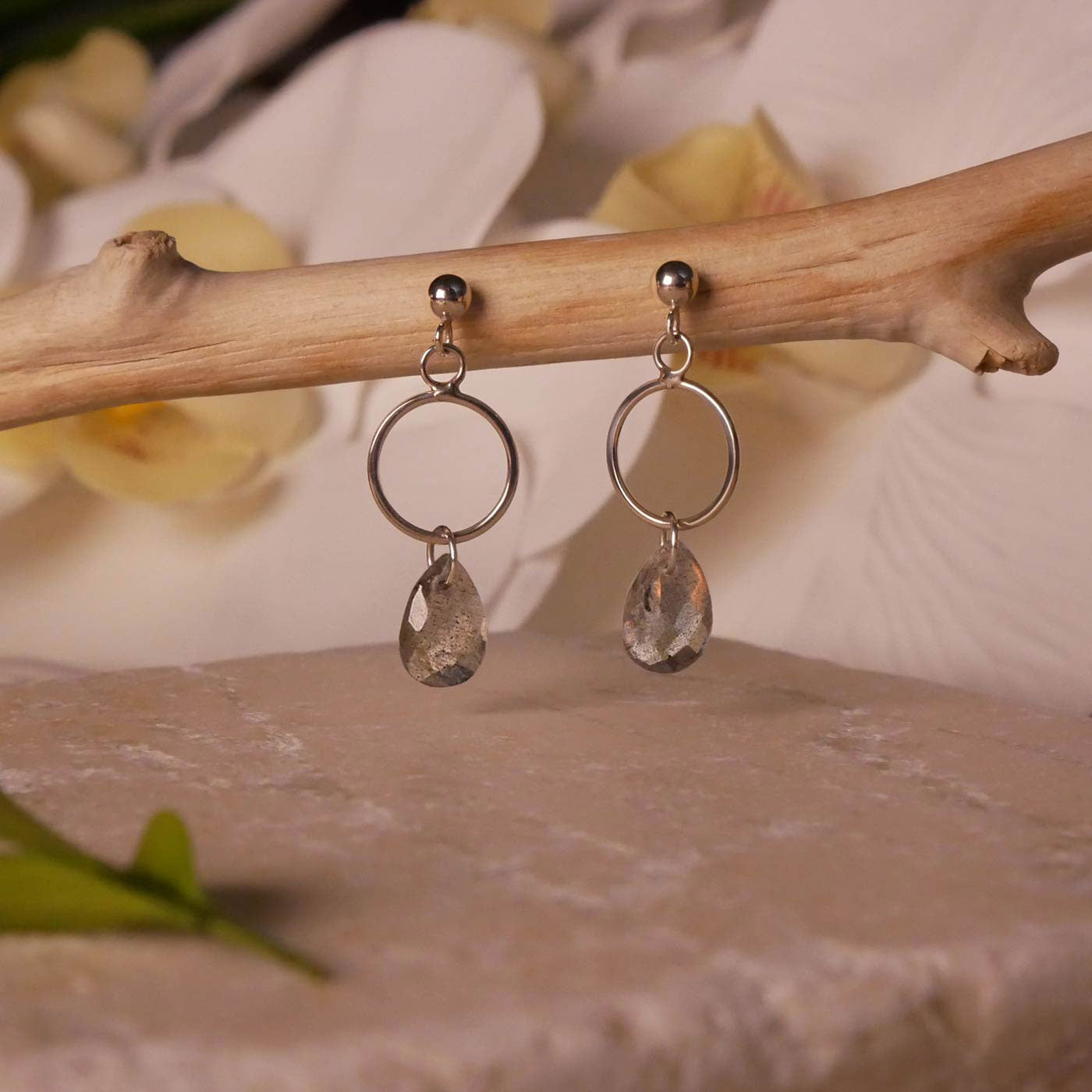 Pair of sterling silver, open circle labradorite drop earrings stylized by dangling on a branch over natural stone surface, with blurred orchid flowers in the background.