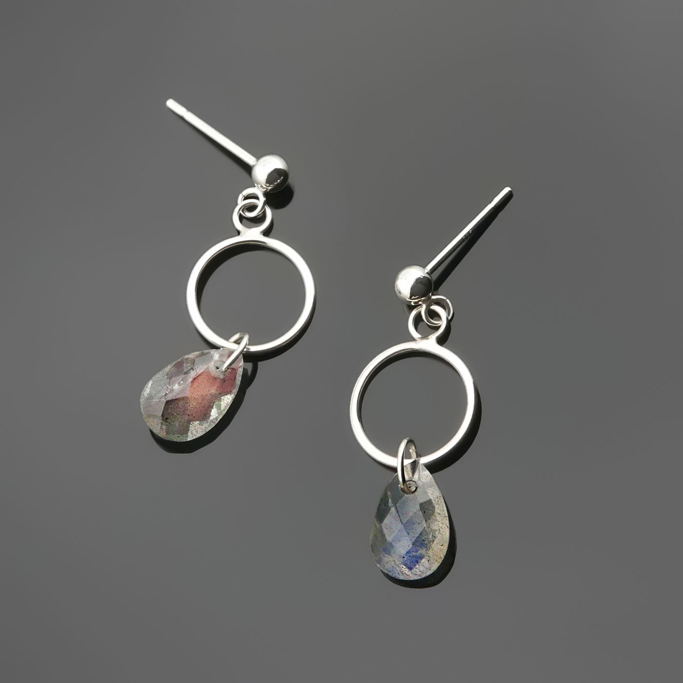 A pair of sterling silver, open circle labradorite drop earrings laying flat on a plain dark gray background, top view.