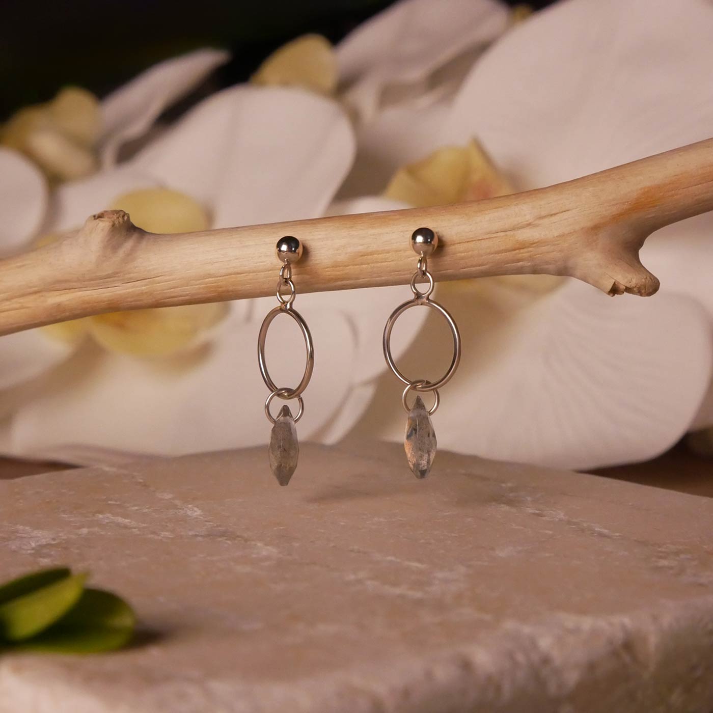 Pair of sterling silver, open circle labradorite drop earrings stylized by dangling on a branch over natural stone surface, with blurred orchid flowers in the background.