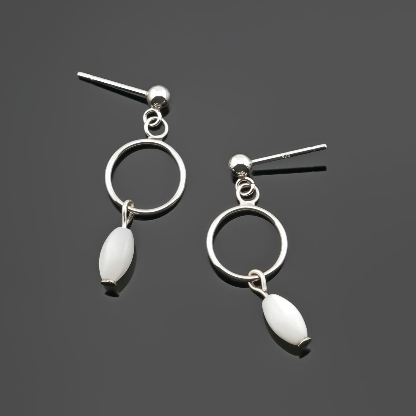 A pair of sterling silver, open circle mother of pearl MOP drop earrings laying flat on a plain dark gray background, top view.