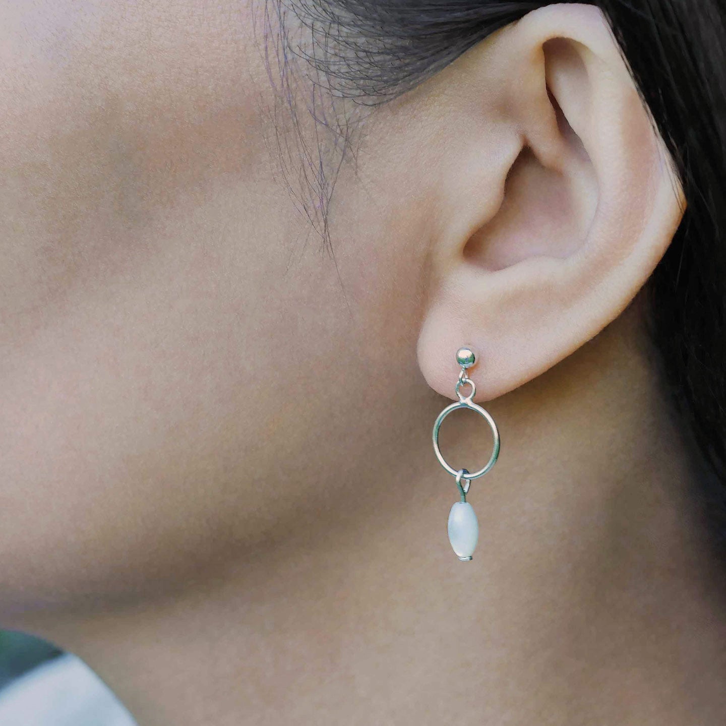 A close up of a sterling silver open circle mother of pearl MOP drop earring worn outside by a person with dark hair.