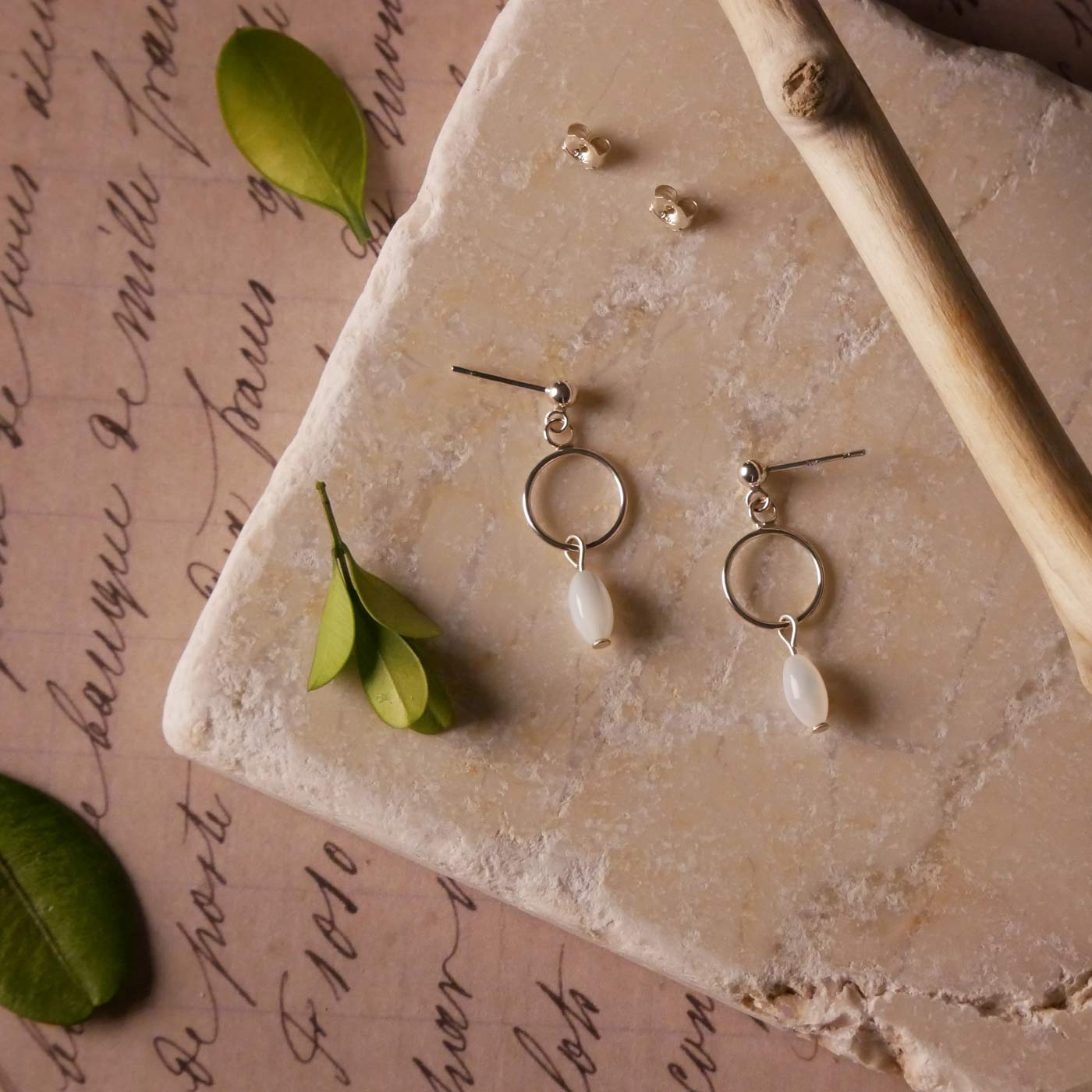 Top view of a pair of sterling silver, open circle mother of pearl MOP drop earrings. It is stylized by laying flat on a natural stone surface, over vintage paper with cursive writing, and surrounded by green leaves with a branch in the foreground.