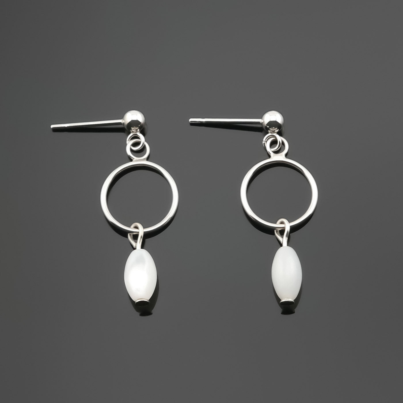 A pair of sterling silver, open circle mother of pearl MOP drop earrings laying flat on a plain dark gray background, top view.