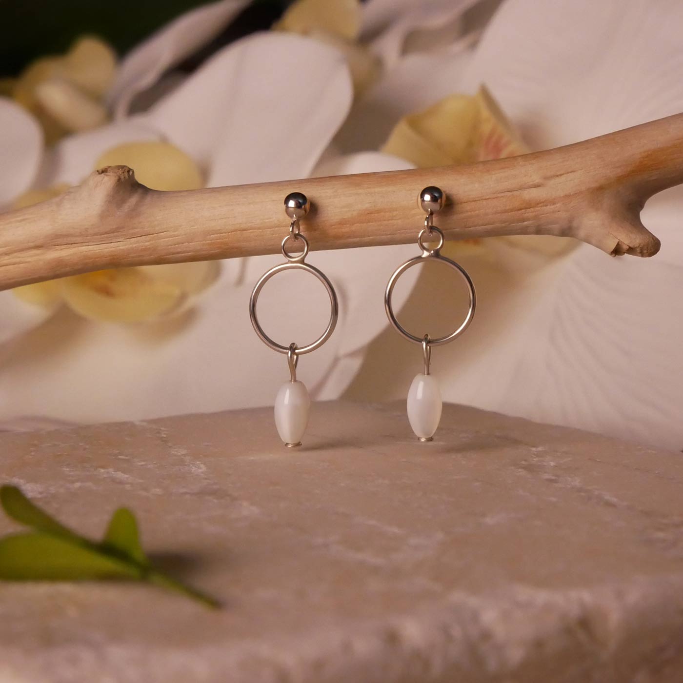 Pair of sterling silver, open circle mother of pearl MOP drop earrings stylized by dangling on a branch over natural stone surface, with blurred orchid flowers in the background.