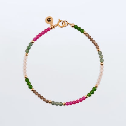 Top view of a dainty multi-colored colorful garden beaded bracelet with natural high quality AAA gemstones of Guatemalan jade, Taiwan jade, lab ruby, smokey quartz and rose quartz with 14K gold filled accent beads, clasp and a reverie blossom branded logo tag laying flat on a plain white or light gray background