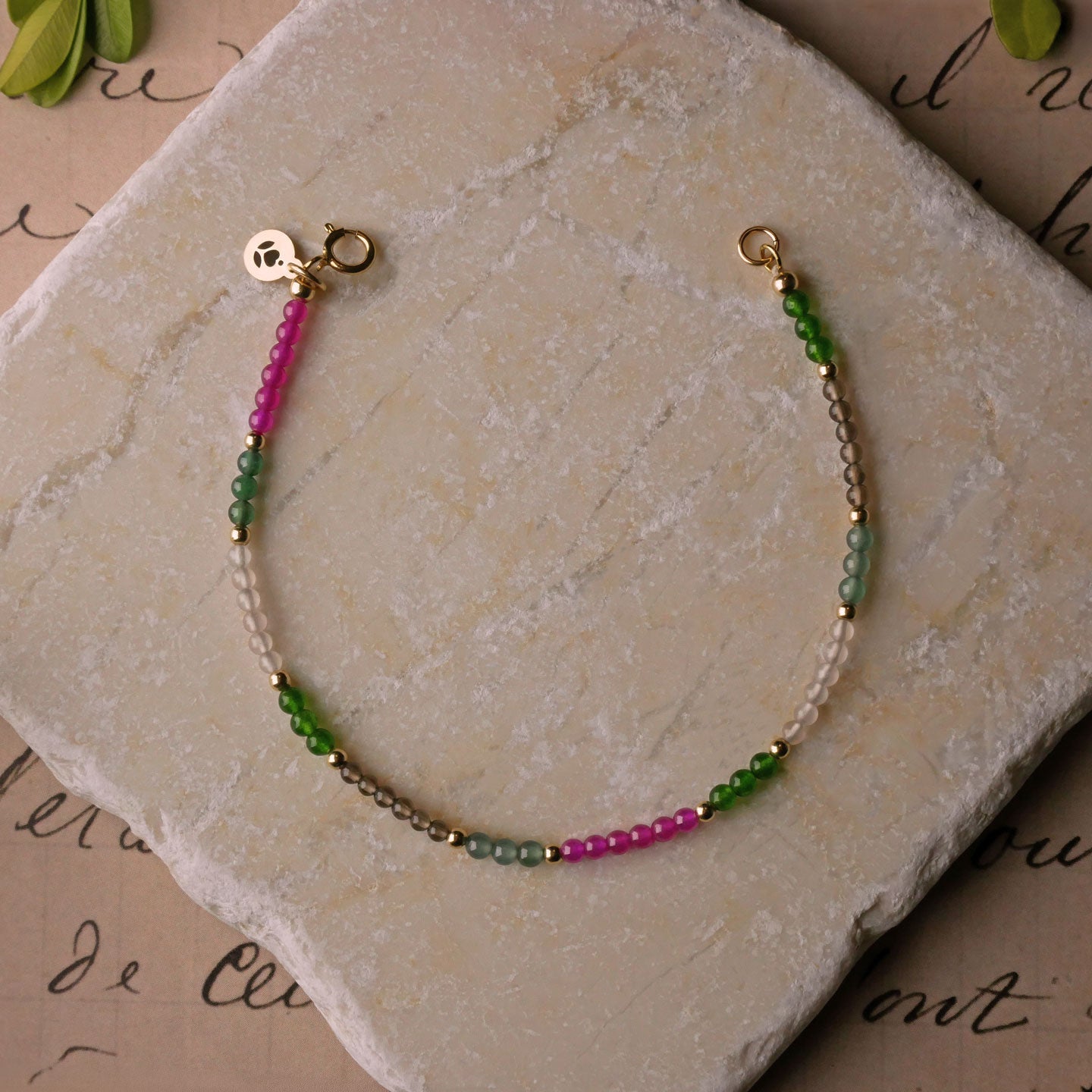 Top view of a dainty multi-colored colorful garden beaded bracelet with natural high quality AAA gemstones of Guatemalan jade, Taiwan jade, lab ruby, smokey quartz and rose quartz with 14K gold filled accent beads, clasp and a reverie blossom branded logo tag laying flat on a stone surface and stylized background with old lettering, vintage light pink beige paper and green leaves.