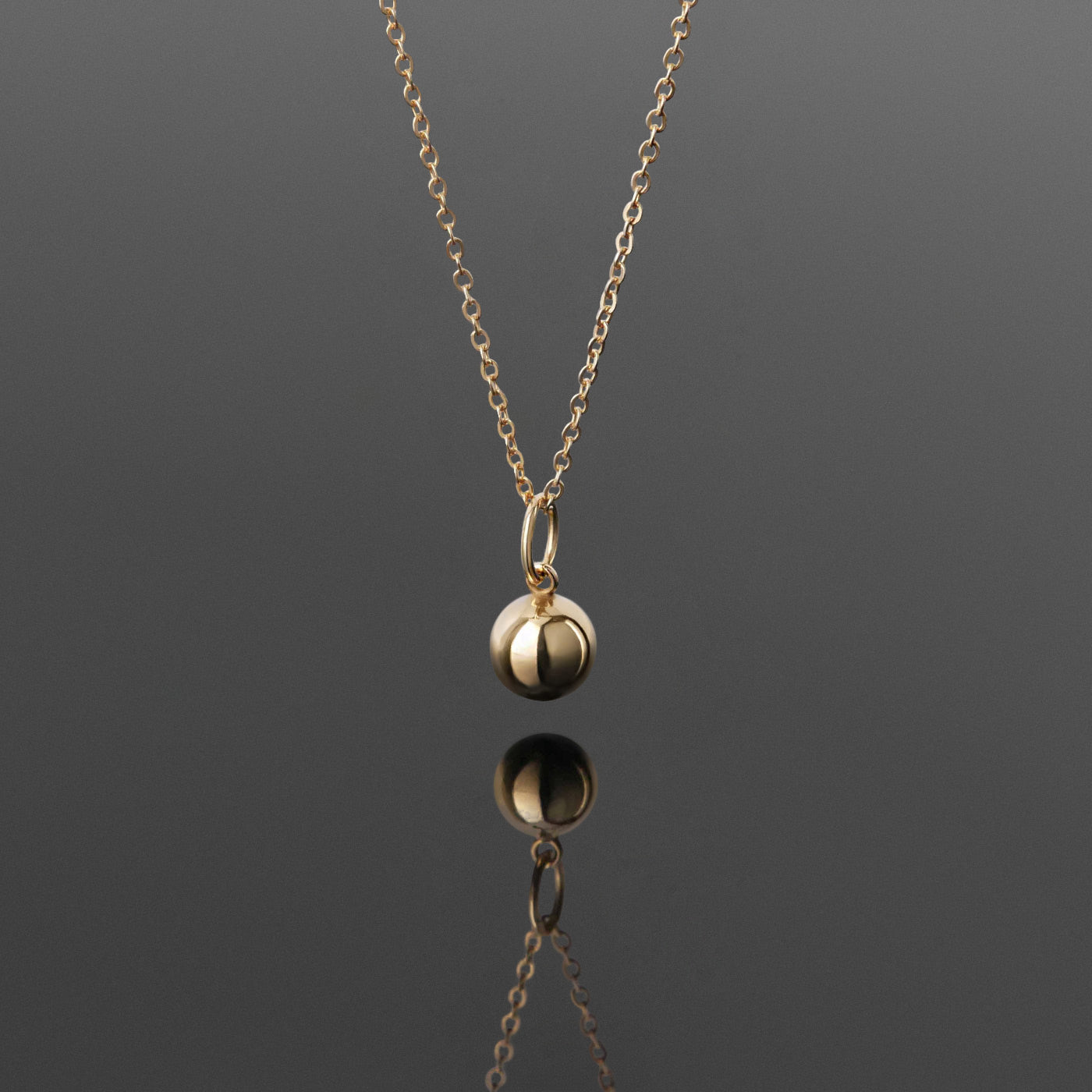 Front view of a dainty, 14k gold filled sphere ball pendant chain necklace hanging, with a plain reflective gray background.