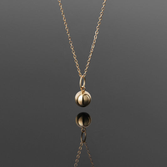 Front view of a dainty, 14k gold filled sphere ball pendant chain necklace hanging, with a plain reflective gray background.