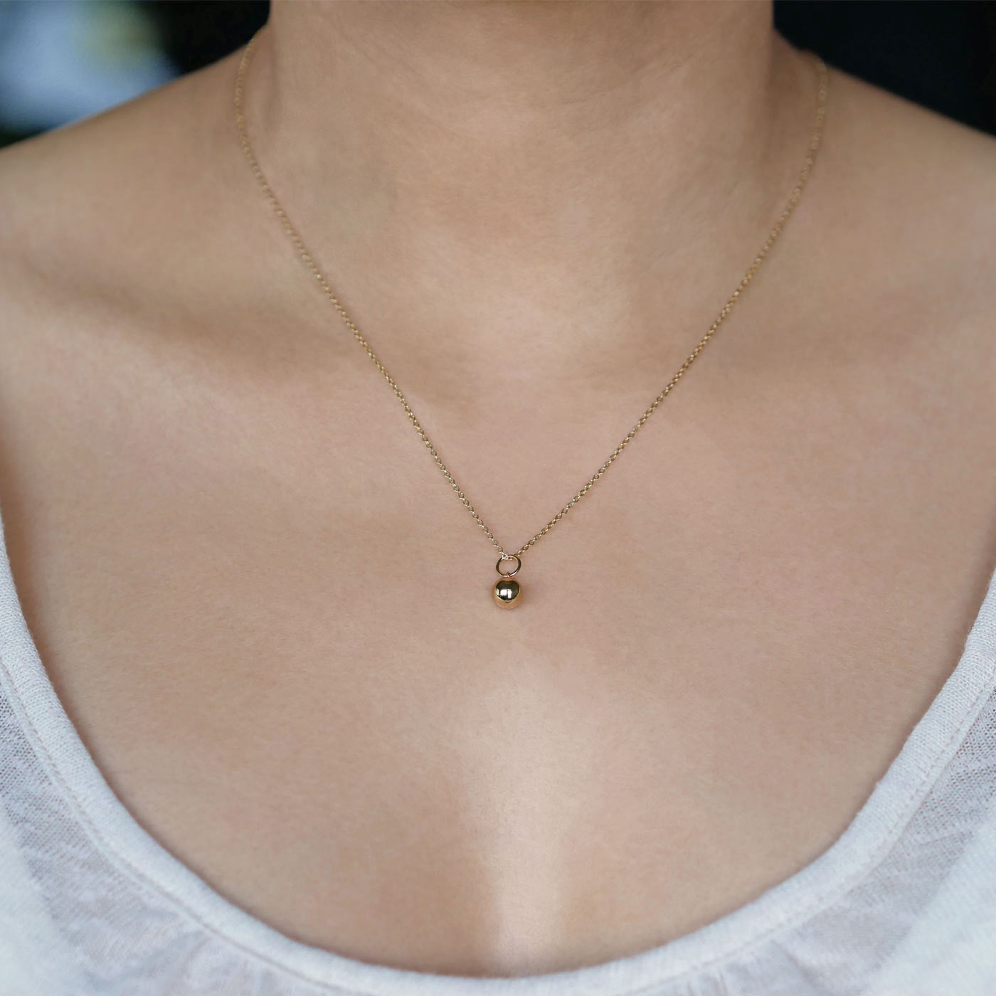 Front view of a dainty, round 14k gold filled sphere ball pendant necklace worn on a person wearing a light beige off white top.
