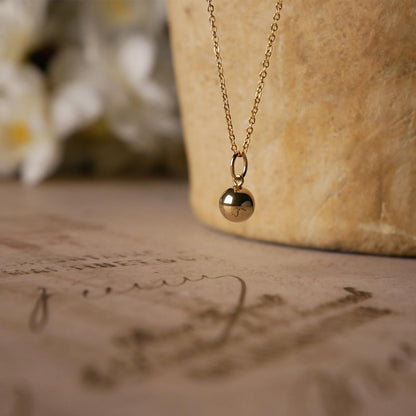 Front side view of a dainty, round 14k gold filled sphere ball pendant necklace. It is stylized by hanging from a textured light brown pot, over vintage paper with old writing style on it, with white flowers blurred in background.