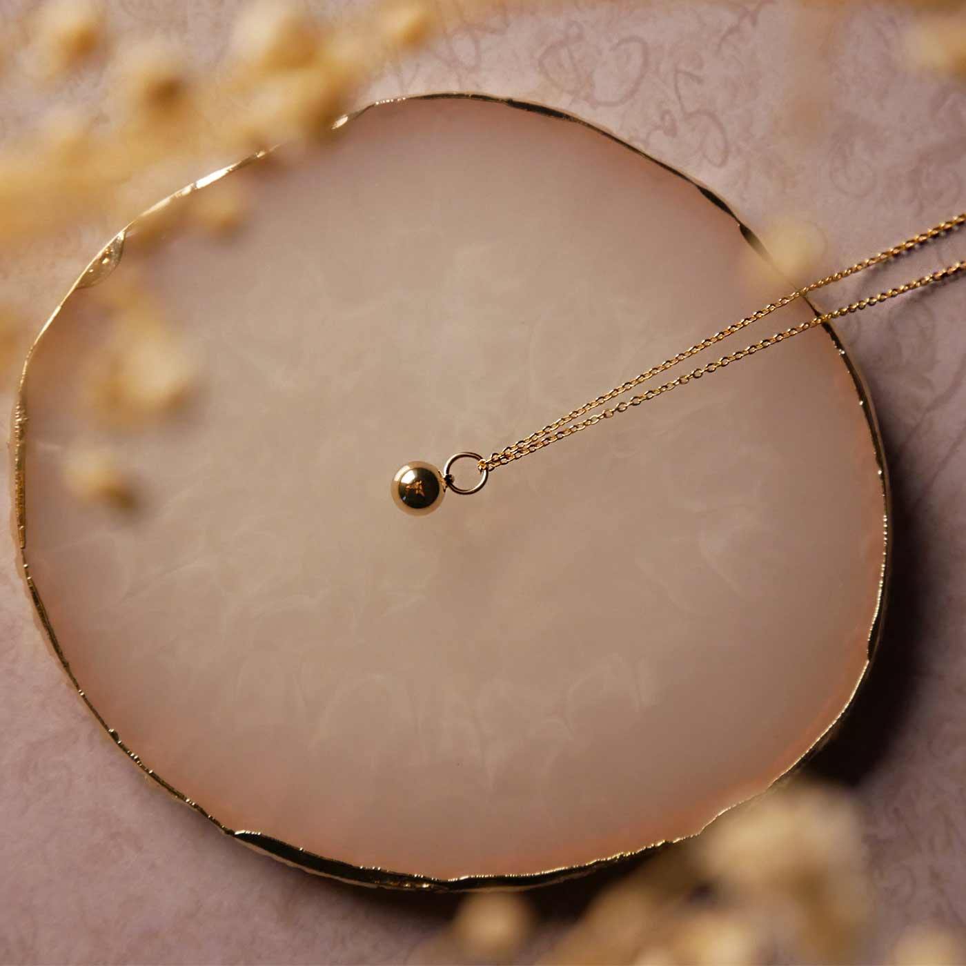 Top view of a dainty, 14k gold filled sphere ball pendant chain necklace laying flat on a stone surface. The stone surface has gold edges, it lays on a vintage patterned beige pink paper. It is also stylized with dried baby's breath small flowers blurred in the foreground.