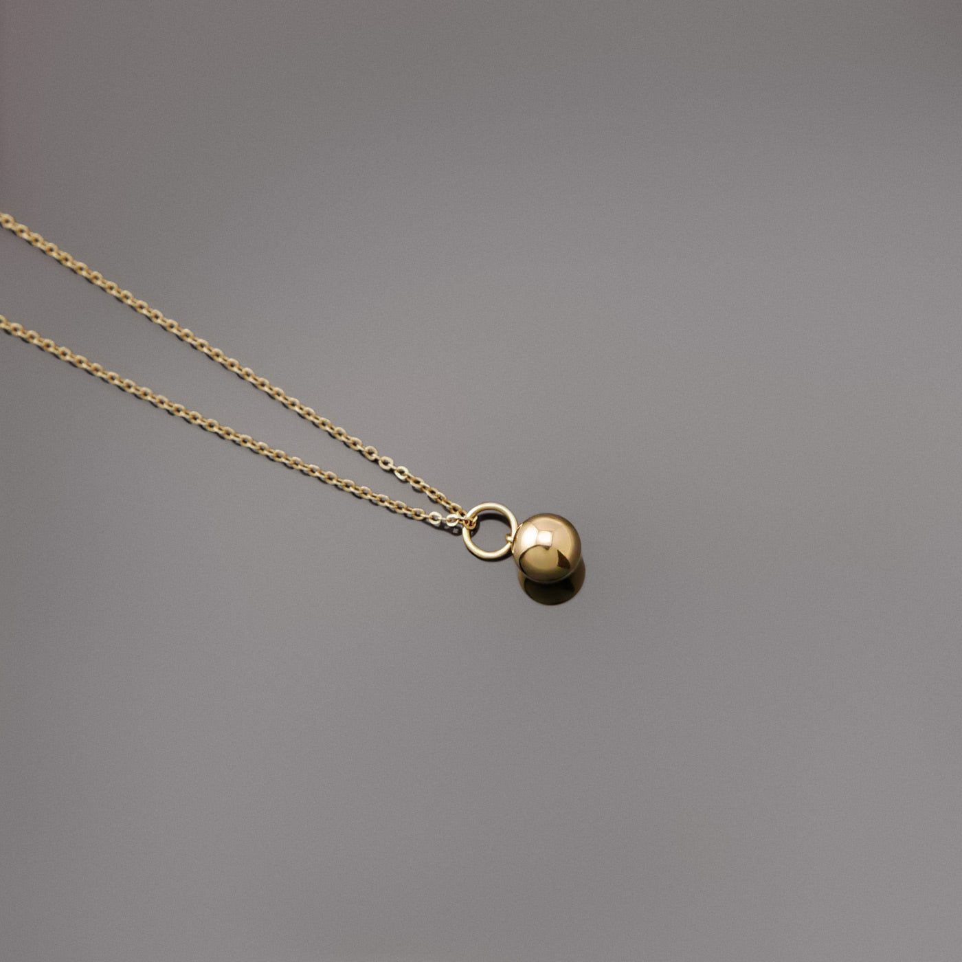 Top view of a dainty, 14k gold filled sphere ball pendant chain necklace laying flat on a plain reflective gray background.