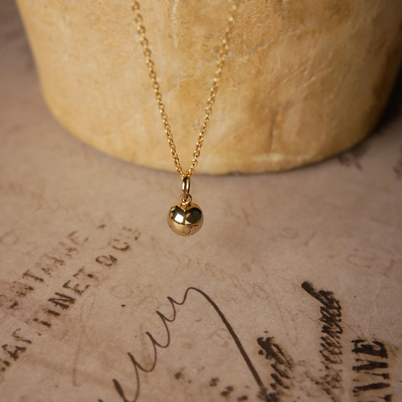 Front view of a dainty, round 14k gold filled sphere ball pendant necklace. It is stylized by hanging from a textured light brown pot, over vintage paper with old writing style on it.