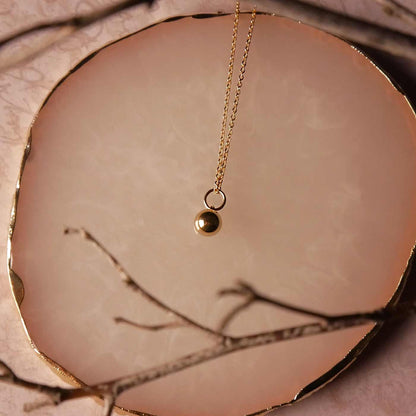 Top view of a dainty, 14k gold filled sphere ball pendant chain necklace laying flat on a stone surface. The stone surface has gold edges, it lays on a vintage patterned beige pink paper. It is also stylized with thin brown branches blurred in the foreground.