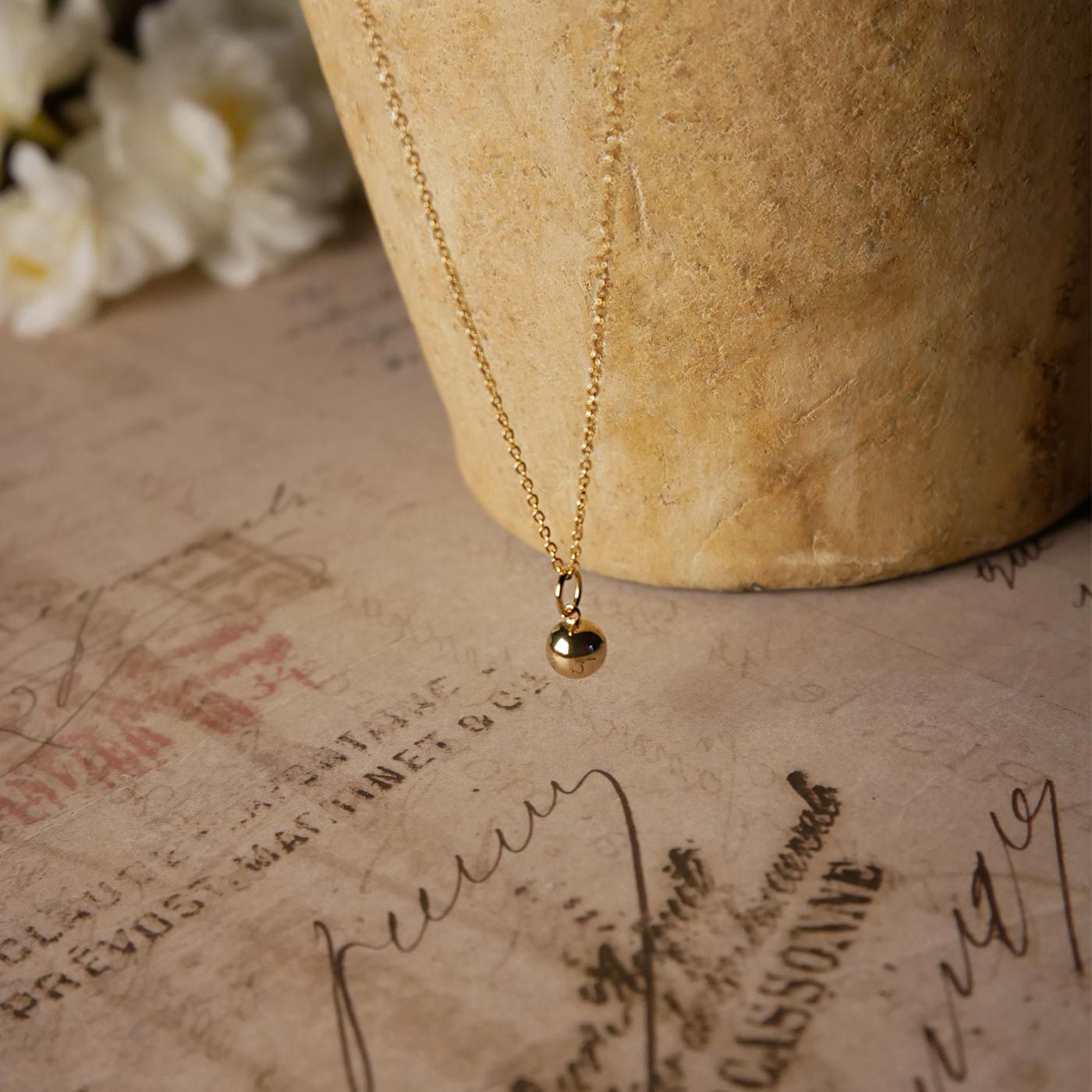 Front top view of a dainty, round 14k gold filled sphere ball pendant necklace. It is stylized by hanging from a textured light brown pot, over vintage paper with old writing style on it, with white flowers blurred in background.