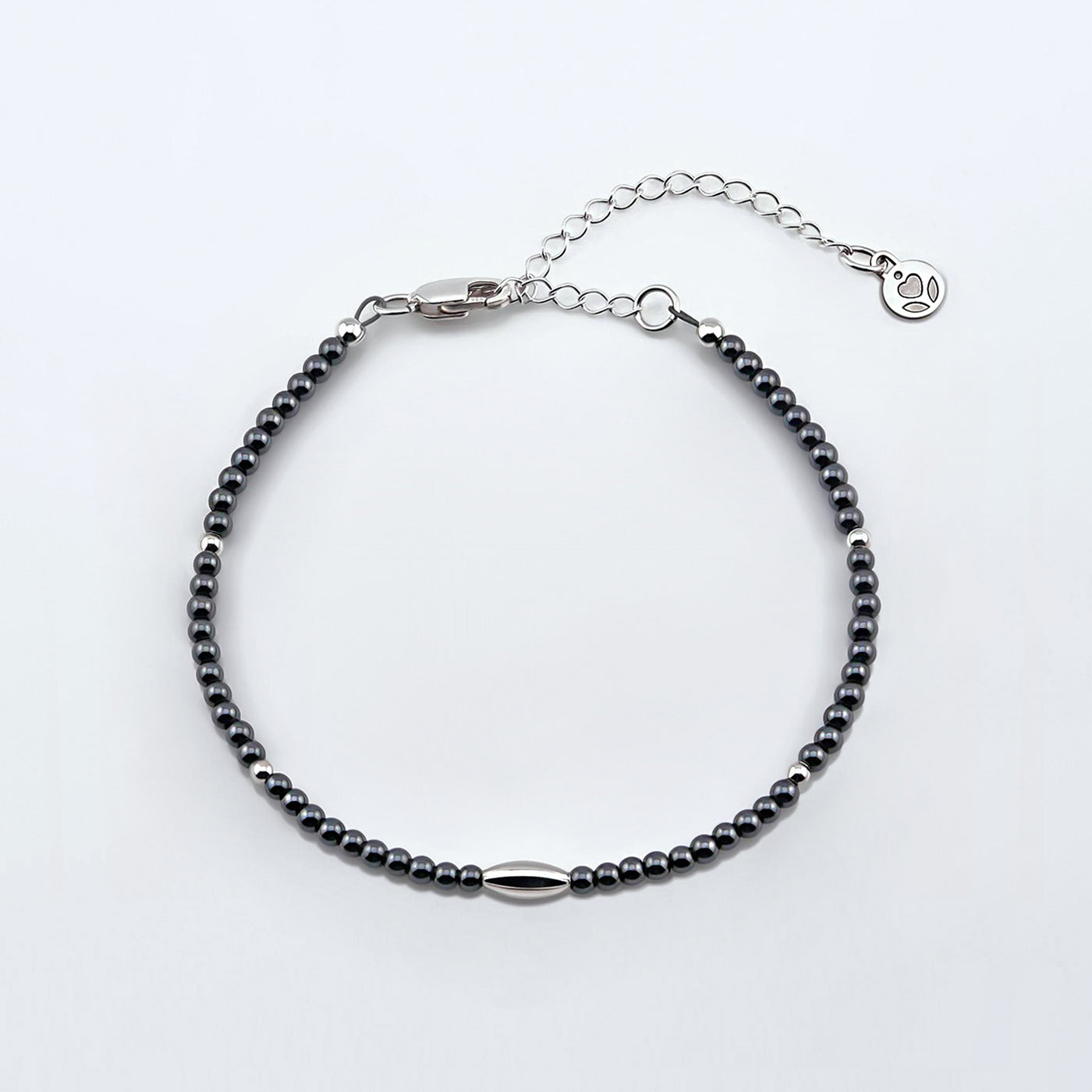 Top view of a dainty gunmetal gray high quality AAA hematite smooth round beaded bracelet with sterling silver focal and accent beads, clasp, reverie blossom brand logo tag and extender chain laying flat on a plain off white or light gray background. 