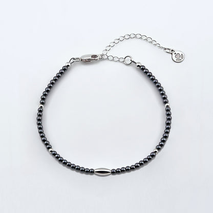 Top view of a dainty gunmetal gray high quality AAA hematite smooth round beaded bracelet with sterling silver focal and accent beads, clasp, reverie blossom brand logo tag and extender chain laying flat on a plain off white or light gray background. 