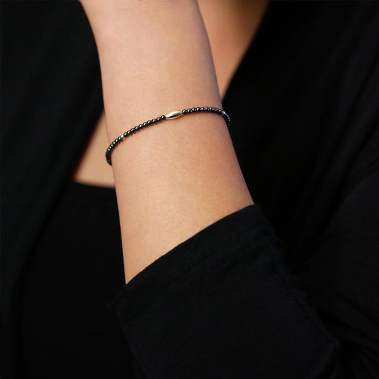 Close-up of a wrist wearing a dainty gunmetal dark gray high quality AAA hematite smooth round beaded bracelet with sterling silver focal and  accent beads. The background is a person wearing a black long sleeve professional style top.