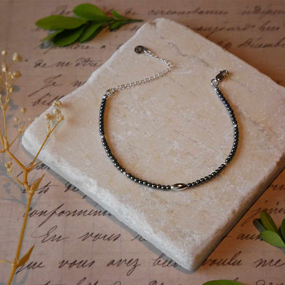 Top front view of a dainty gunmetal gray high quality AAA hematite smooth round beaded bracelet with sterling silver accent beads, clasp, reverie blossom brand logo tag and extender chain. It is laying flat on a stone surface stylized with a background of vintage light pink beige paper underneath with old cursive writing, dried beige baby breath flowers and green leaves.
