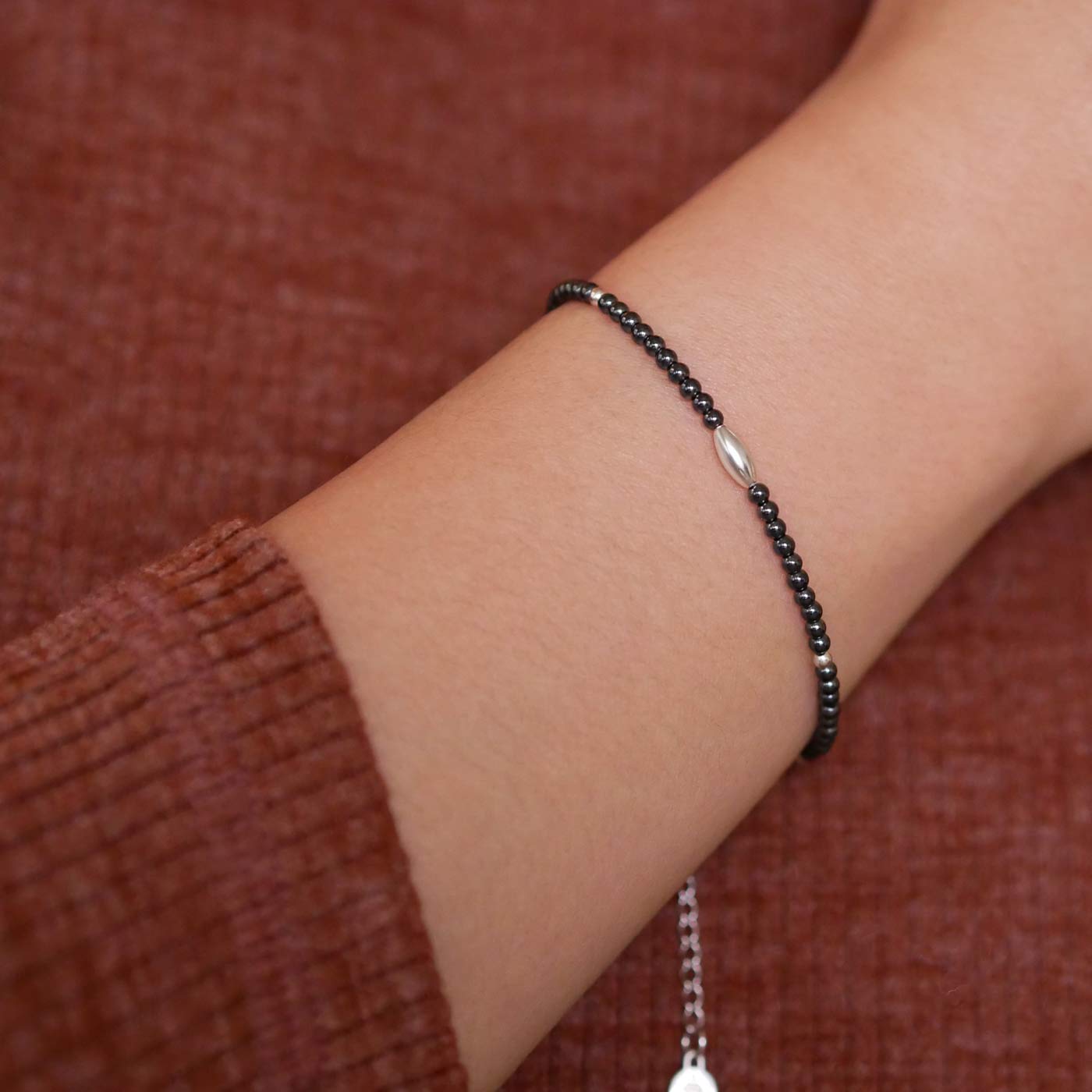 Close-up of a wrist wearing a dainty gunmetal dark gray high quality AAA hematite smooth round beaded bracelet with sterling silver focal and  accent beads. The background is a person wearing a brown long sleeve sweater.