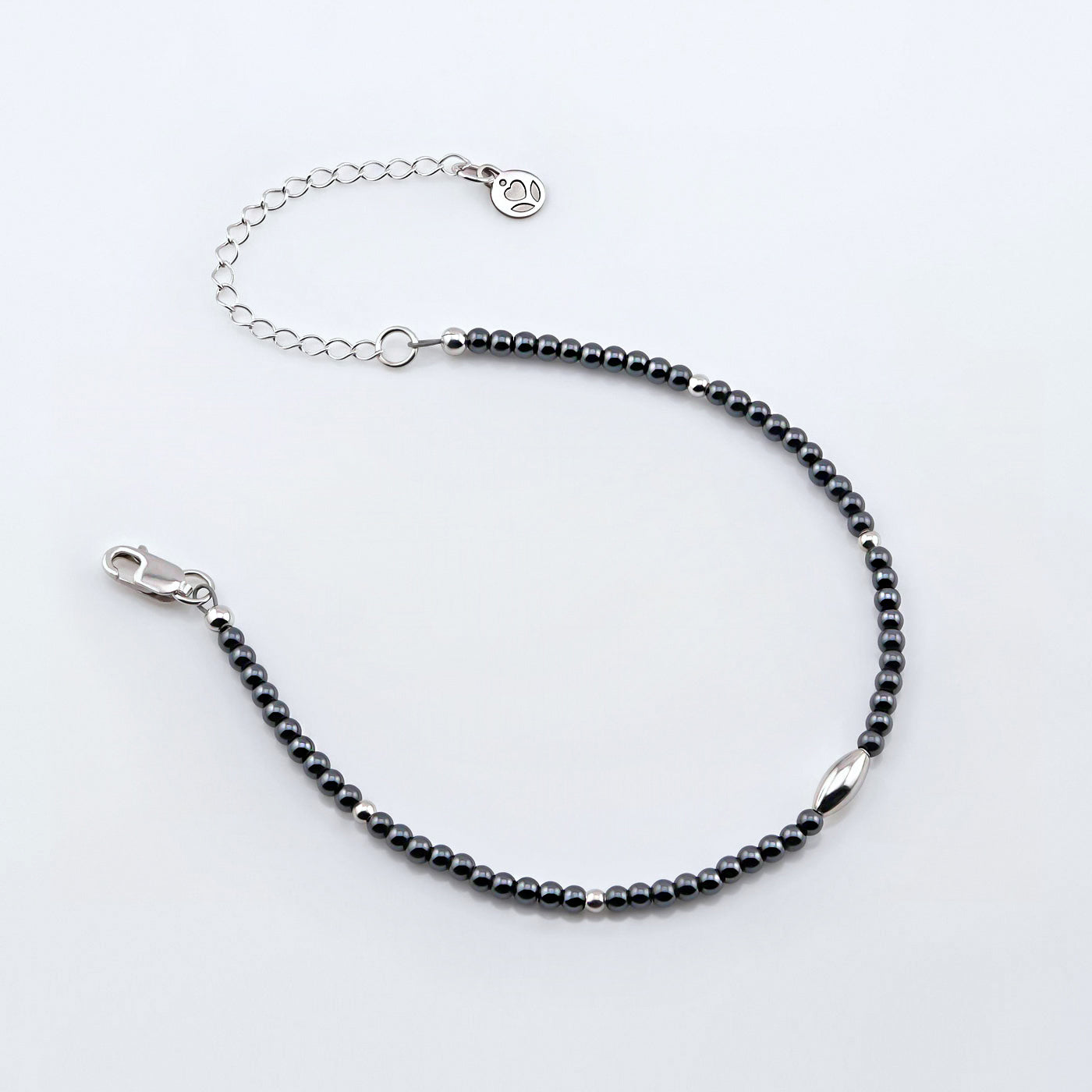 Top angle view of a dainty gunmetal gray high quality AAA hematite smooth round beaded bracelet with sterling silver focal and accent beads, clasp, reverie blossom brand logo tag and extender chain laying flat on a plain off white or light gray background. 