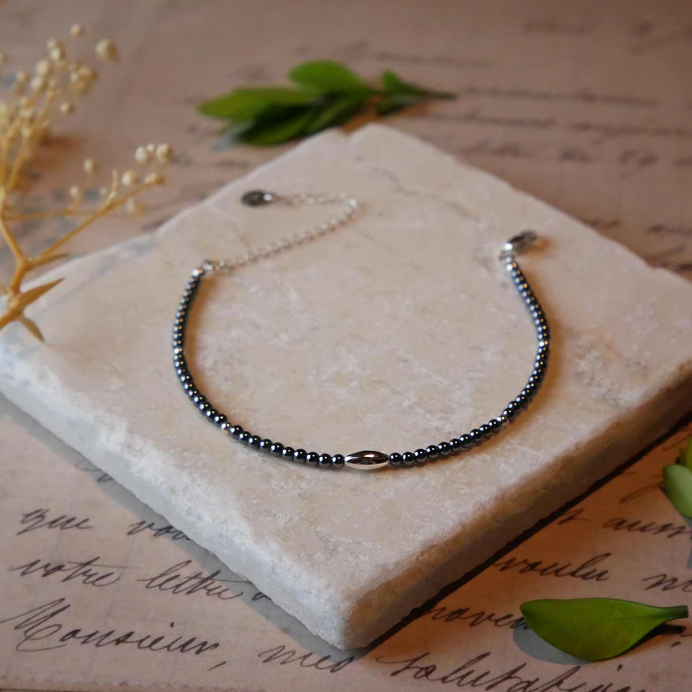 Front view of a dainty gunmetal gray high quality AAA hematite smooth round beaded bracelet with sterling silver focal and accent beads, clasp, reverie blossom brand logo tag and extender chain. It is laying flat on a stone surface stylized with a background of vintage light pink beige paper underneath with old cursive writing, dried beige baby breath flowers and green leaves.