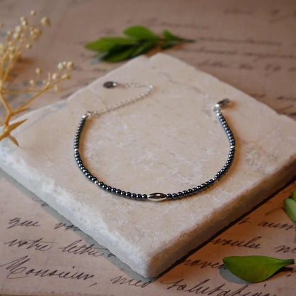 Front view of a dainty gunmetal gray high quality AAA hematite smooth round beaded bracelet with sterling silver focal and accent beads, clasp, reverie blossom brand logo tag and extender chain. It is laying flat on a stone surface stylized with a background of vintage light pink beige paper underneath with old cursive writing, dried beige baby breath flowers and green leaves.