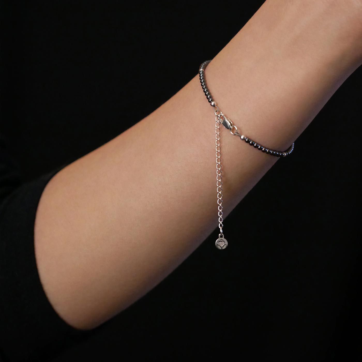 Close-up of a wrist wearing a dainty gunmetal dark gray high quality AAA hematite smooth round beaded bracelet with sterling silver accent beads, clasp, reverie blossom brand logo and extender chain on a black background.