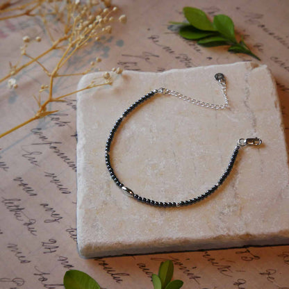 Top 3/4 view of a dainty gunmetal gray high quality AAA hematite smooth round beaded bracelet with sterling silver focal and accent beads, clasp, reverie blossom brand logo tag and extender chain. It is laying flat on a stone surface stylized with a background of vintage light pink beige paper underneath with old cursive writing, dried beige baby breath flowers and green leaves.