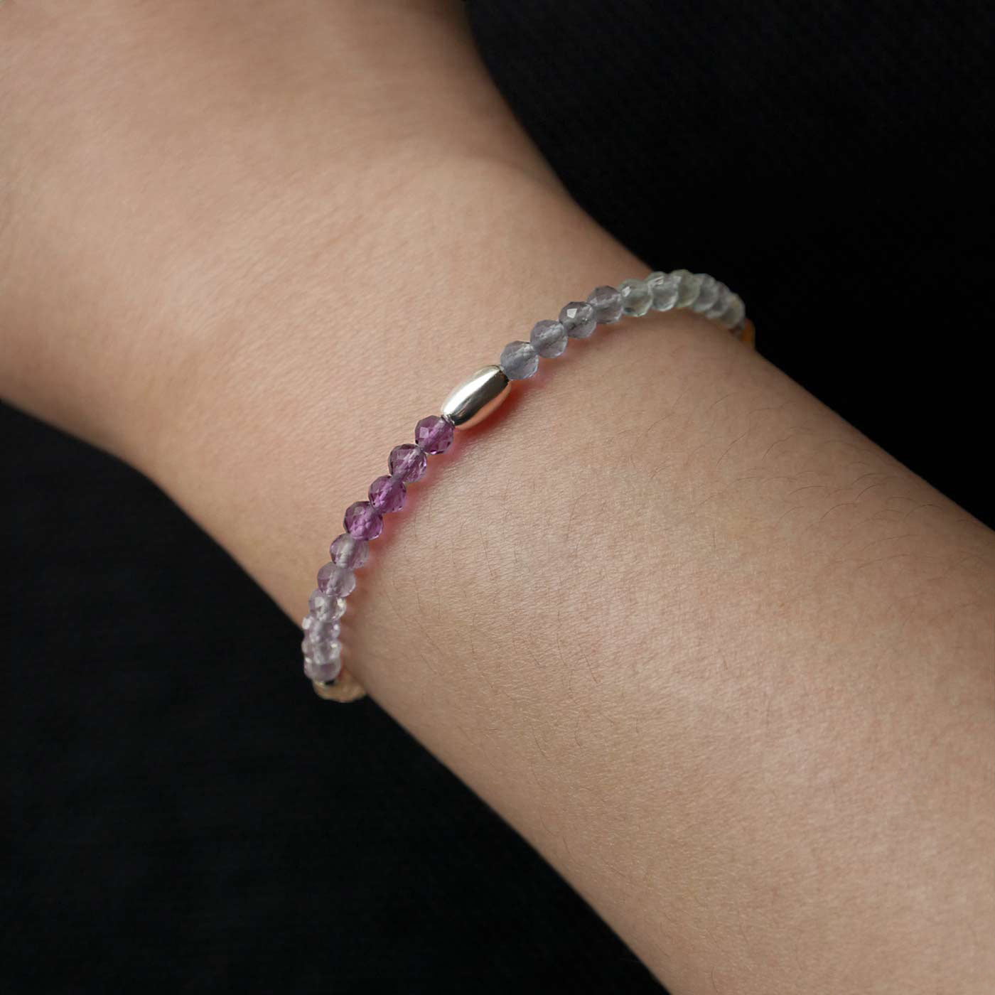 Close-up of a wrist wearing a multi-colored, colorful, ombre high quality AAA rainbow fluorite faceted beaded bracelet with sterling silver accent beads, on a dark black background.