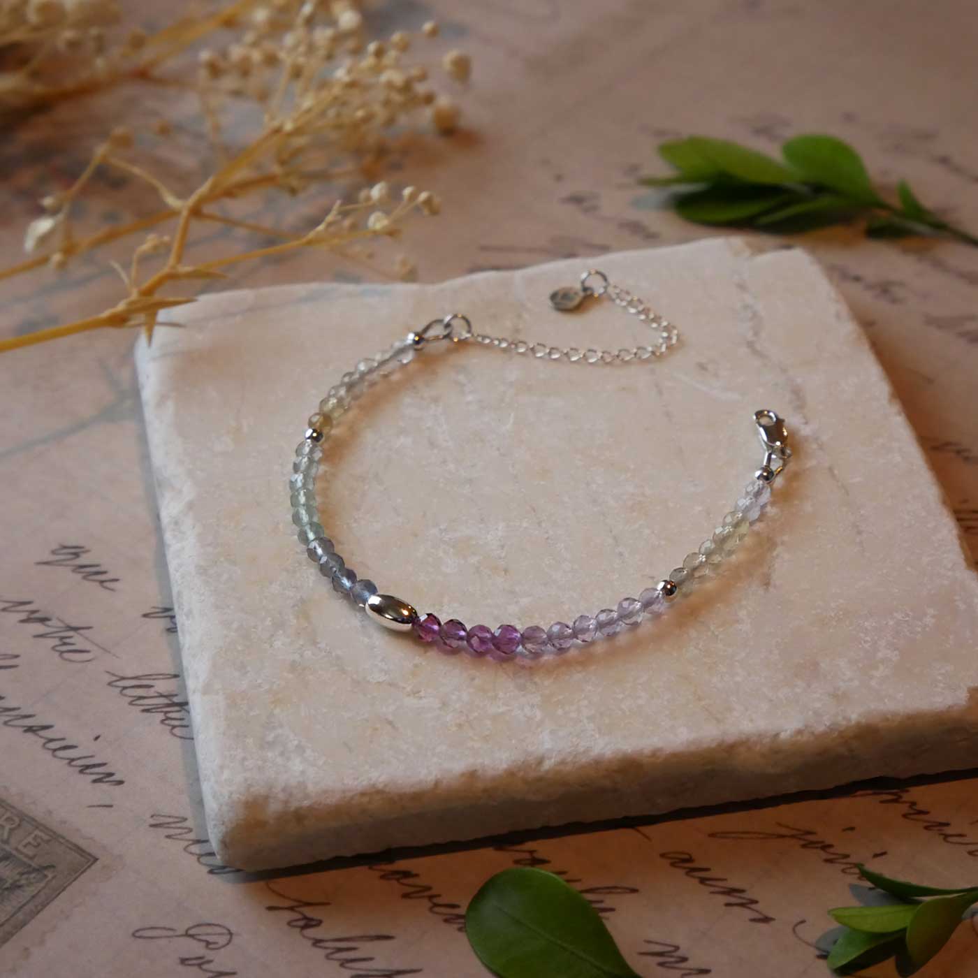 3/4 view of a dainty multi-colored, colorful ombre high quality AAA rainbow fluorite faceted beaded bracelet with sterling silver accent beads, clasp, reverie blossom branded logo tag and extender chain. It is laying flat on a stone surface stylized with a background of vintage light pink beige paper underneath with old lettering, dried beige baby breath flowers and green leaves.