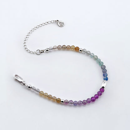 Top view of a multi-colored, colorful ombre high quality AAA rainbow fluorite faceted beaded bracelet with sterling silver accent beads, clasp, reverie blossom brand logo and extender chain laying flat on a plain off white or light gray background. 