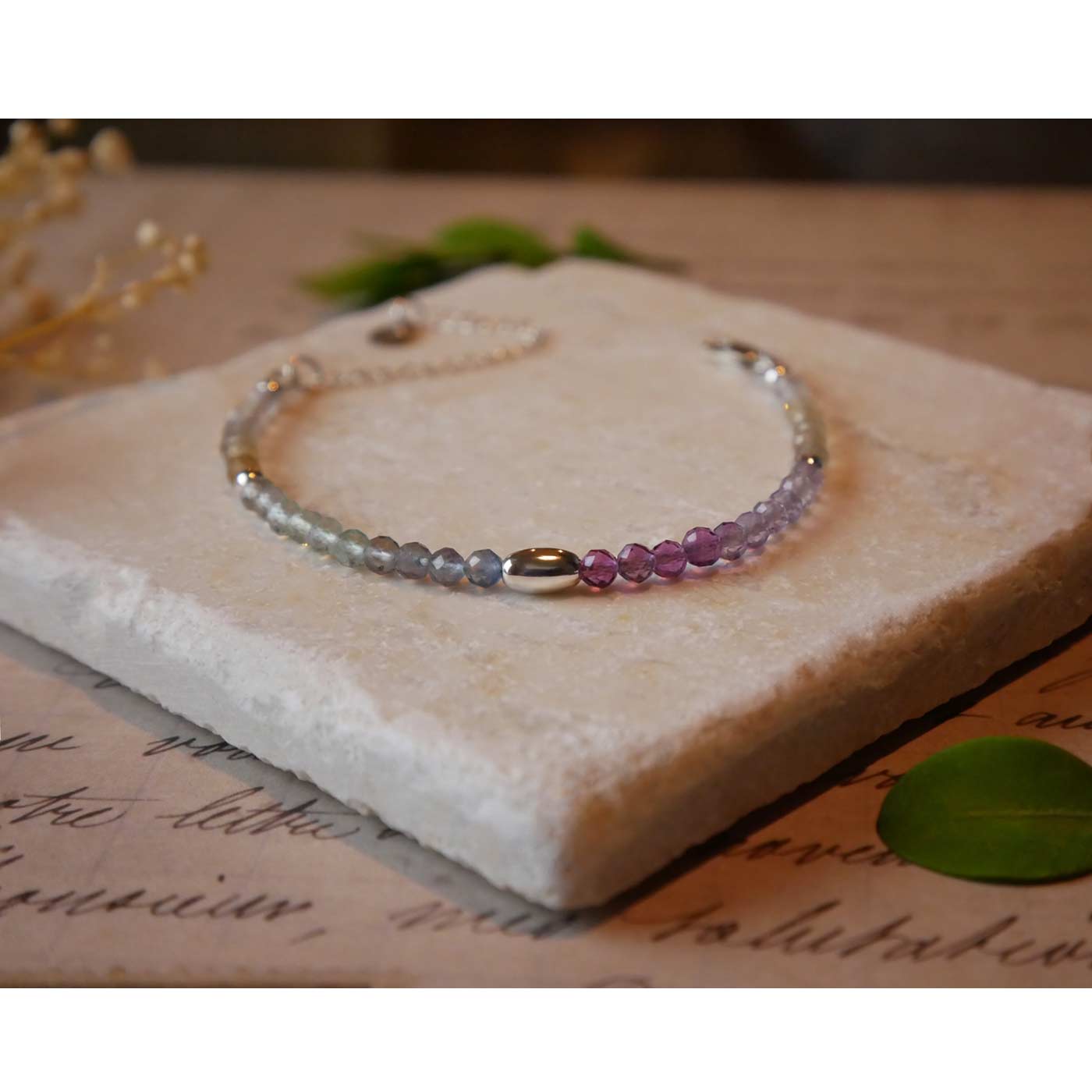 Close up front view of a dainty multi-colored, colorful ombre high quality AAA rainbow fluorite faceted beaded bracelet with sterling silver accent beads, clasp, reverie blossom branded logo tag and extender chain. It is laying flat on a stone surface stylized with a background of vintage light pink beige paper underneath with old cursive writing, dried beige baby breath flowers and green leaves.