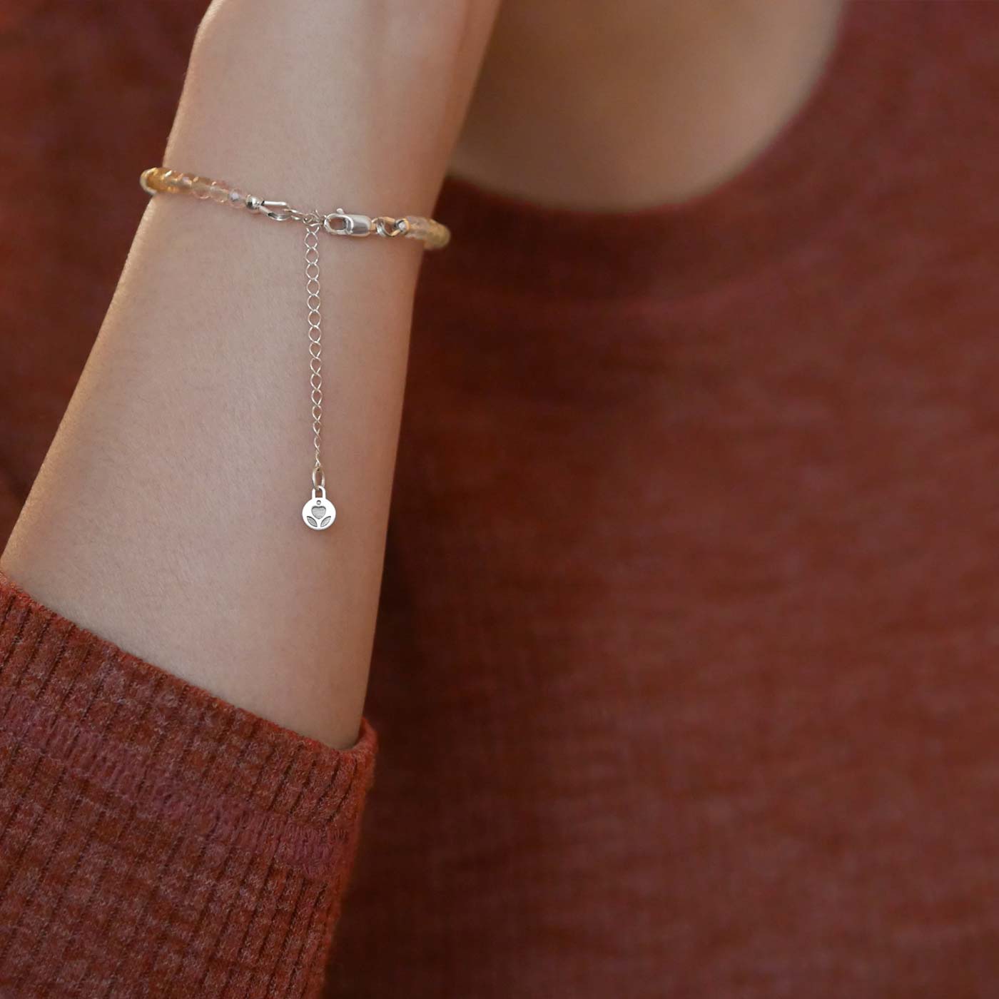 Close up of a person wearing a brown long sleeve shirt, wearing on their wrist a multi-colored, colorful ombre high quality AAA rainbow fluorite faceted beaded bracelet with sterling silver accent beads, clasp, reverie blossom brand logo and extender chain hanging.