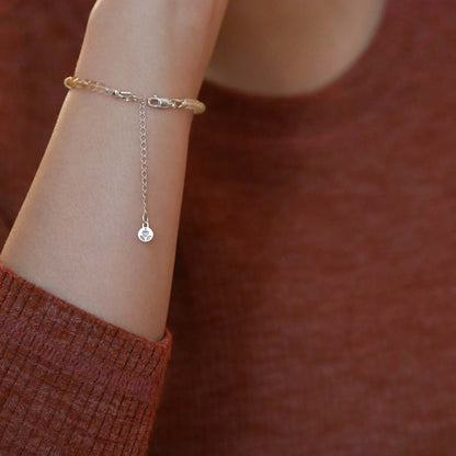 Close up of a person wearing a brown long sleeve shirt, wearing on their wrist a multi-colored, colorful ombre high quality AAA rainbow fluorite faceted beaded bracelet with sterling silver accent beads, clasp, reverie blossom brand logo and extender chain hanging.