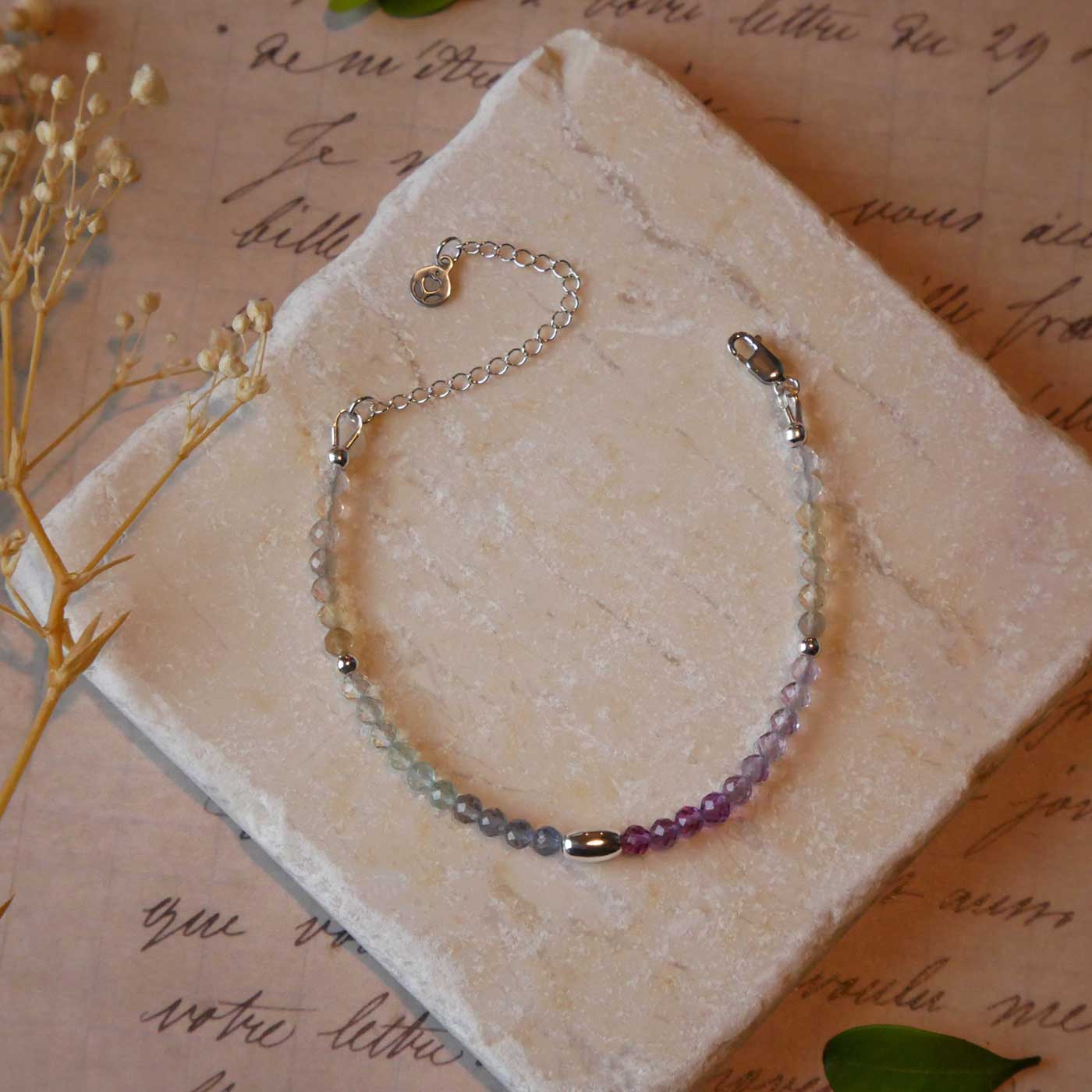 Top view of a dainty multi-colored, colorful ombre high quality AAA rainbow fluorite faceted beaded bracelet with sterling silver accent beads, clasp, reverie blossom branded logo tag and extender chain. It is laying flat on a stone surface stylized with a background of vintage light pink beige paper underneath with old cursive writing, dried beige baby breath flowers and green leaves.