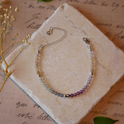 Top view of a dainty multi-colored, colorful ombre high quality AAA rainbow fluorite faceted beaded bracelet with sterling silver accent beads, clasp, reverie blossom branded logo tag and extender chain. It is laying flat on a stone surface stylized with a background of vintage light pink beige paper underneath with old cursive writing, dried beige baby breath flowers and green leaves.
